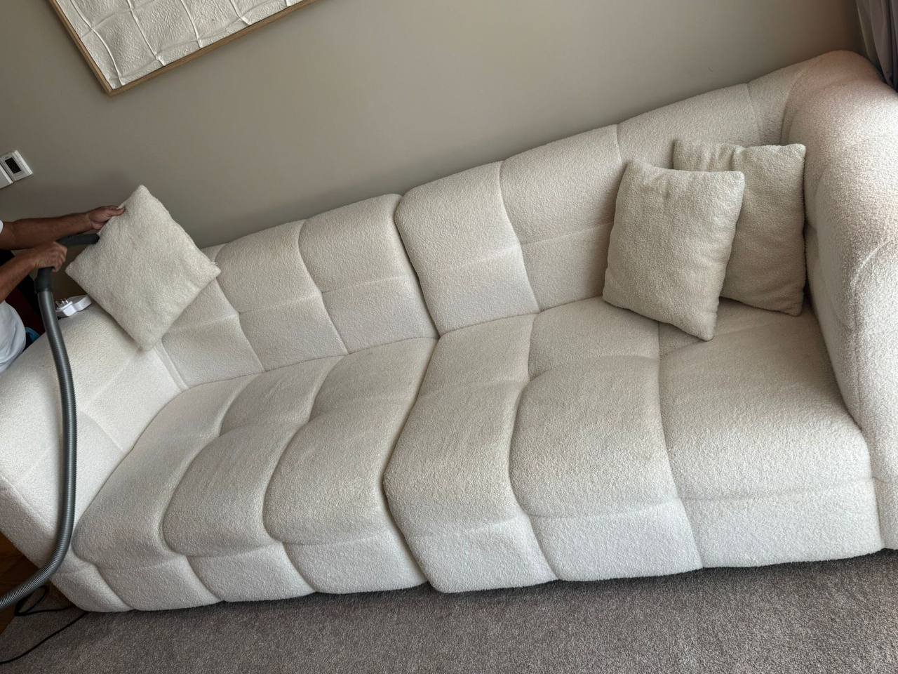 Person cleaning a beige sofa with a vacuum cleaner. The sofa has a tufted design and there are two beige throw pillows on