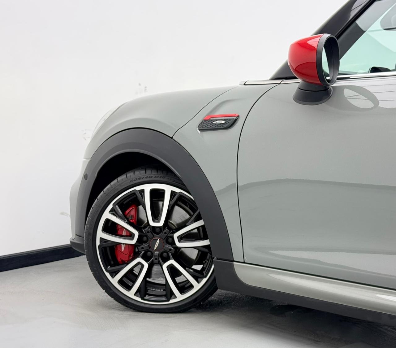 Side view of a grey sports car. The car has a black roof and a red taillight on the right side. The front wheel of the car