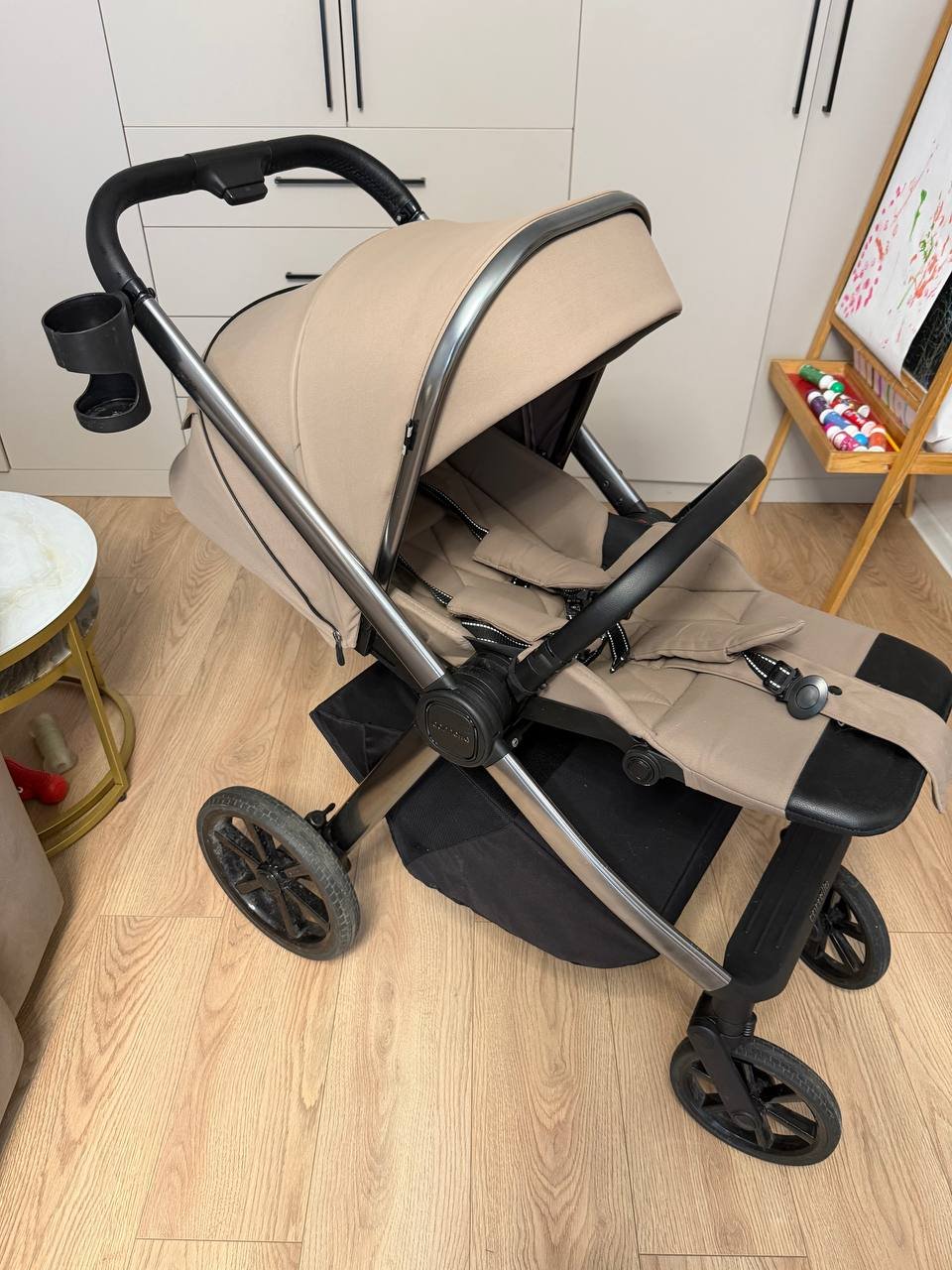 Baby stroller in a room with wooden flooring. The stroller is beige in color and has a black handlebar and seat. It has four