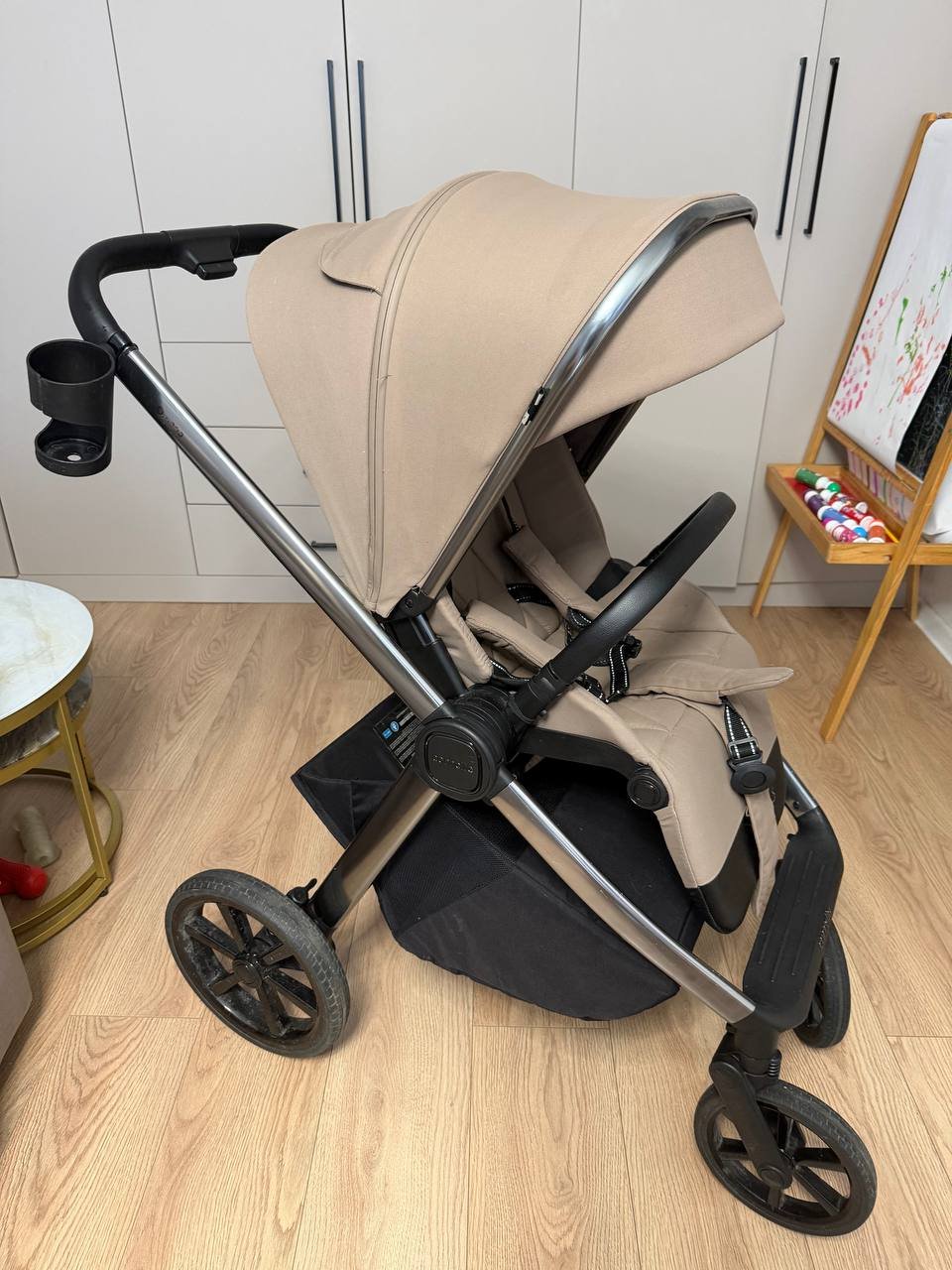 Baby stroller in a room with wooden flooring. The stroller is beige in color and has a black handlebar and wheels. It has a