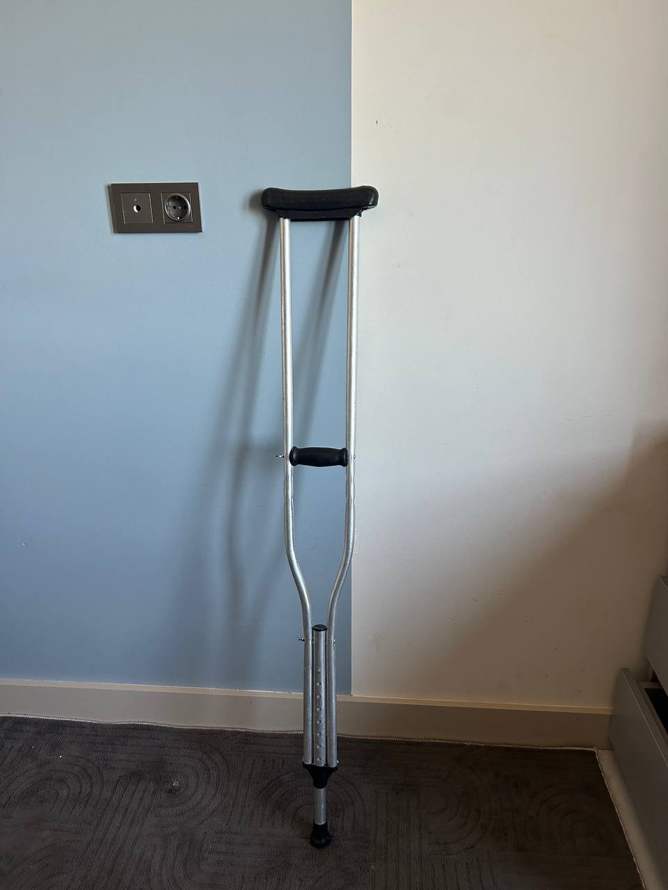 Crutch leaning against a blue wall in a corner of a room. The crutch is made of metal and has a black handle and footrest.