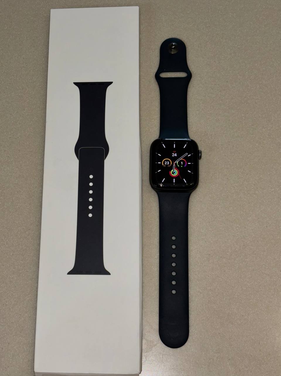 Apple Watch in its packaging box. The box is white with a black strap on the left side. The watch has a round face with a