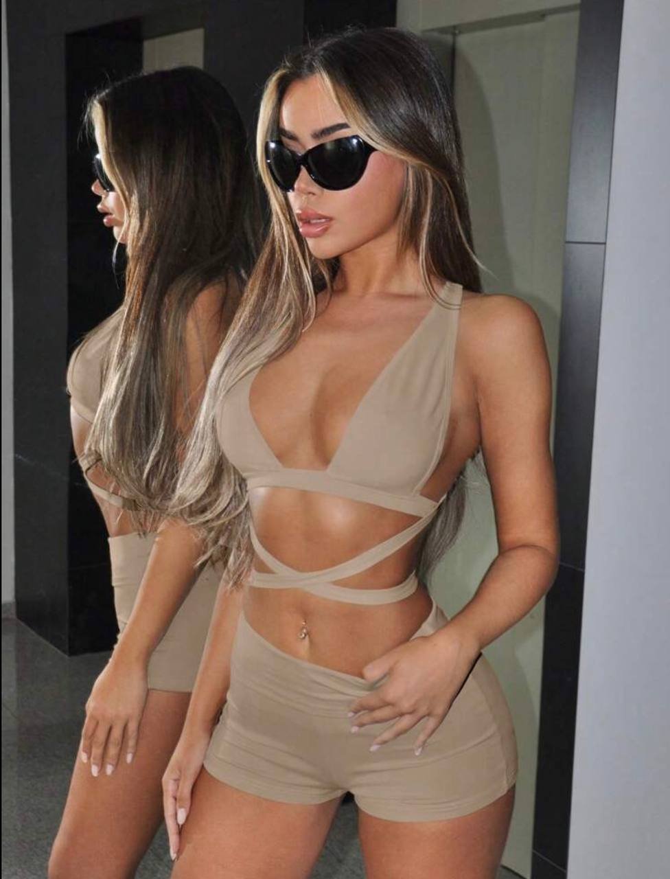 Young woman standing in front of a mirror, wearing a beige two-piece swimsuit. The swimsuit has a deep V-neckline with thin