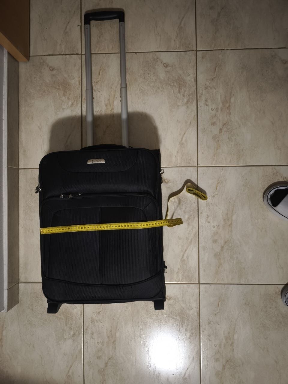 Black suitcase with a yellow measuring tape wrapped around it. The suitcase is lying on a tiled floor and there is a pair of