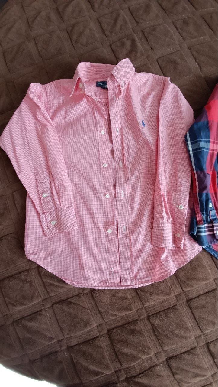 Pink button-down shirt lying on a brown quilted bedspread. The shirt has a collar and long sleeves with a small blue logo on
