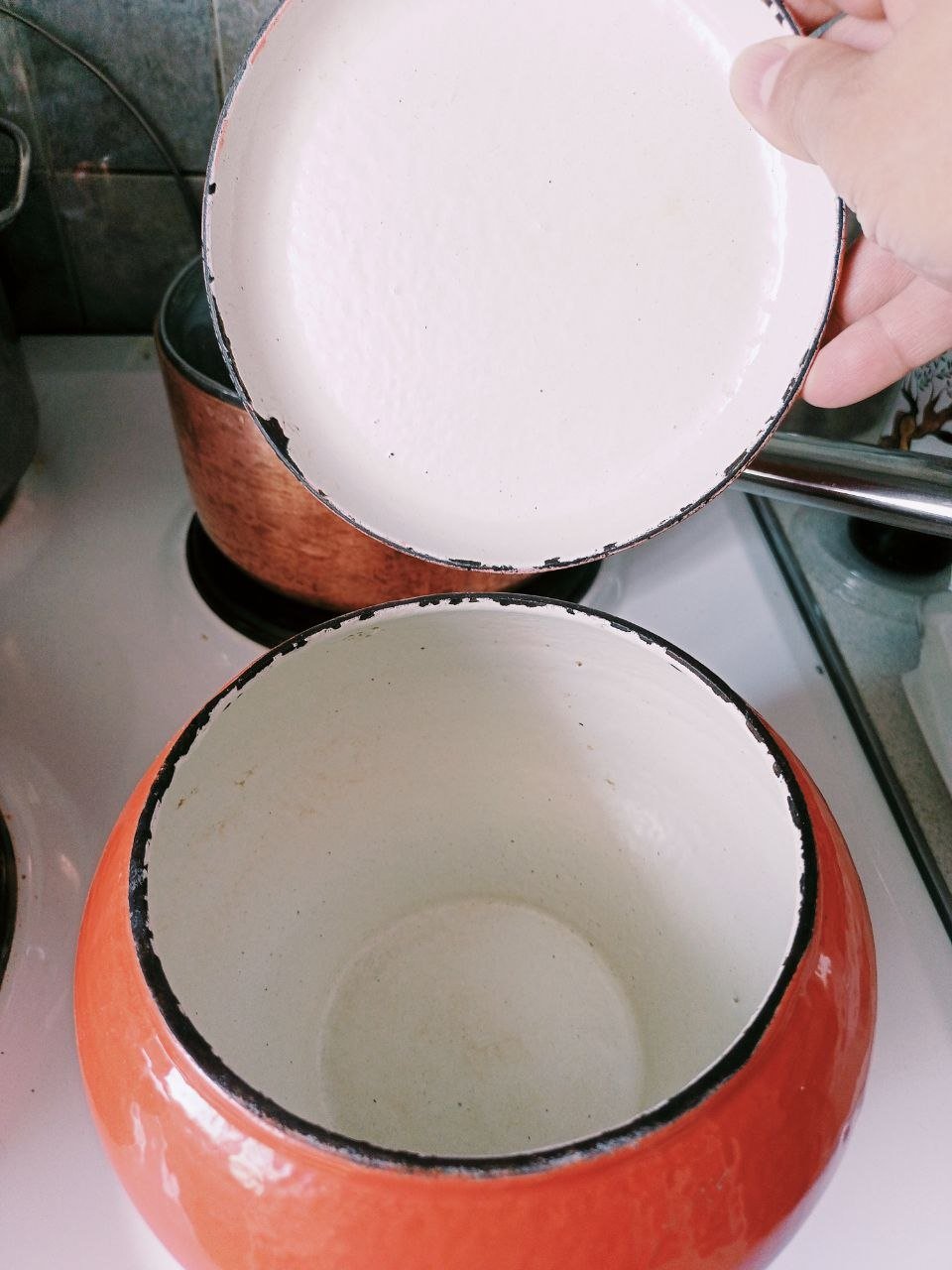 Person's hand holding a white frying pan over an orange ceramic pot on a gas stove. The pot appears to be empty and is