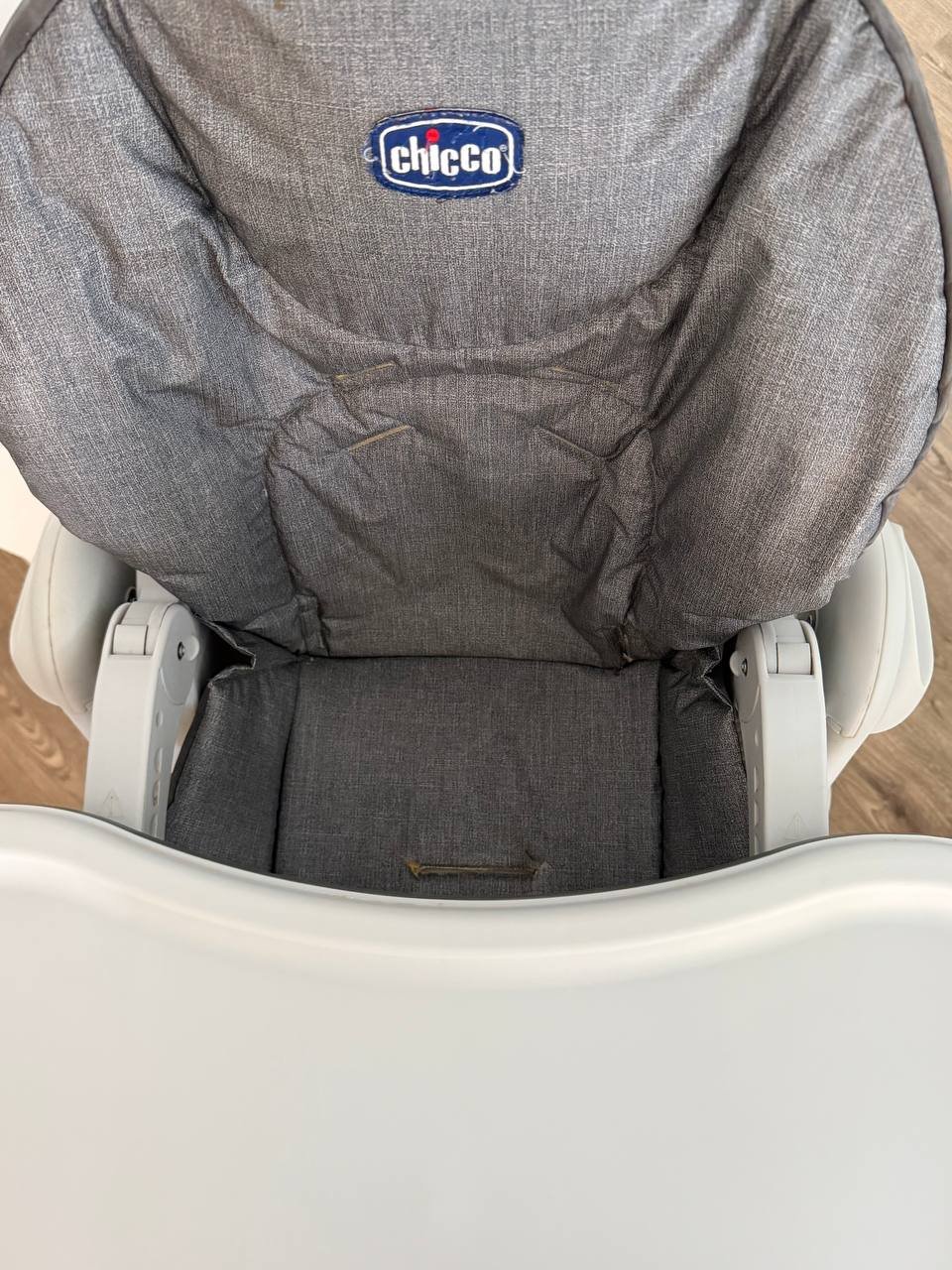 Back of a Chicco high chair. The chair is white and has a grey fabric seat cover with the Chicco logo in blue and red. The