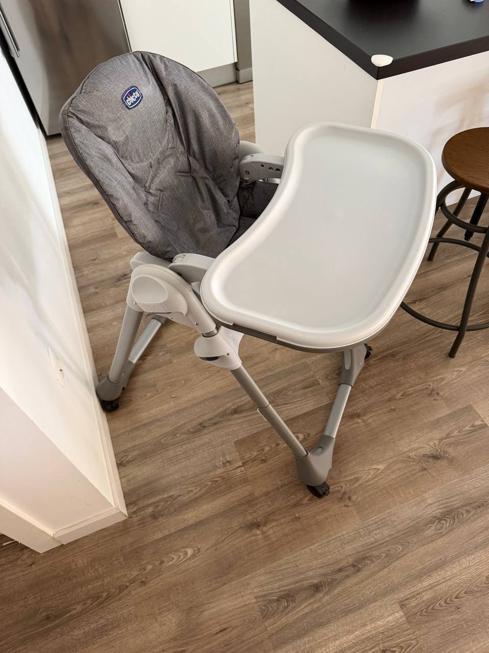 White high chair in a kitchen. The chair has a grey seat and backrest with a blue logo on it. It is attached to a white