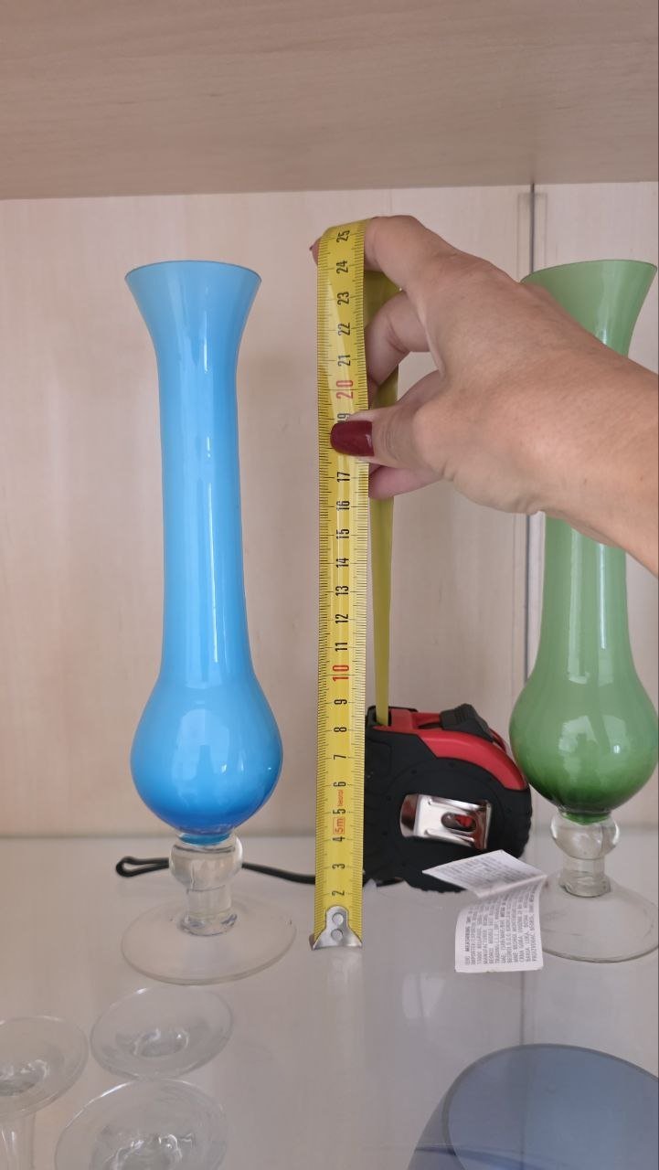 Person's hand holding a yellow measuring tape and measuring a blue vase. The vase is on a white surface and there are two