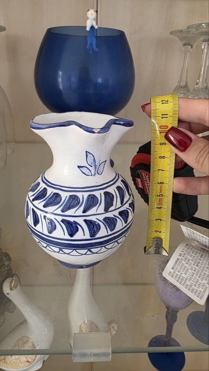 Hand holding a yellow measuring tape over a white ceramic vase. The vase has a blue and white design with a leaf pattern on