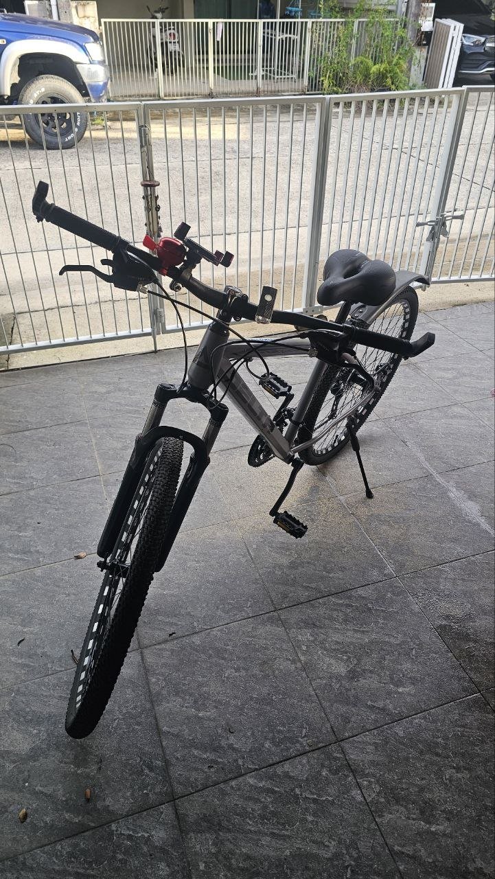 Black bicycle leaning against a metal fence on a sidewalk. The bicycle has a black seat and handlebars, and a red handlebar.