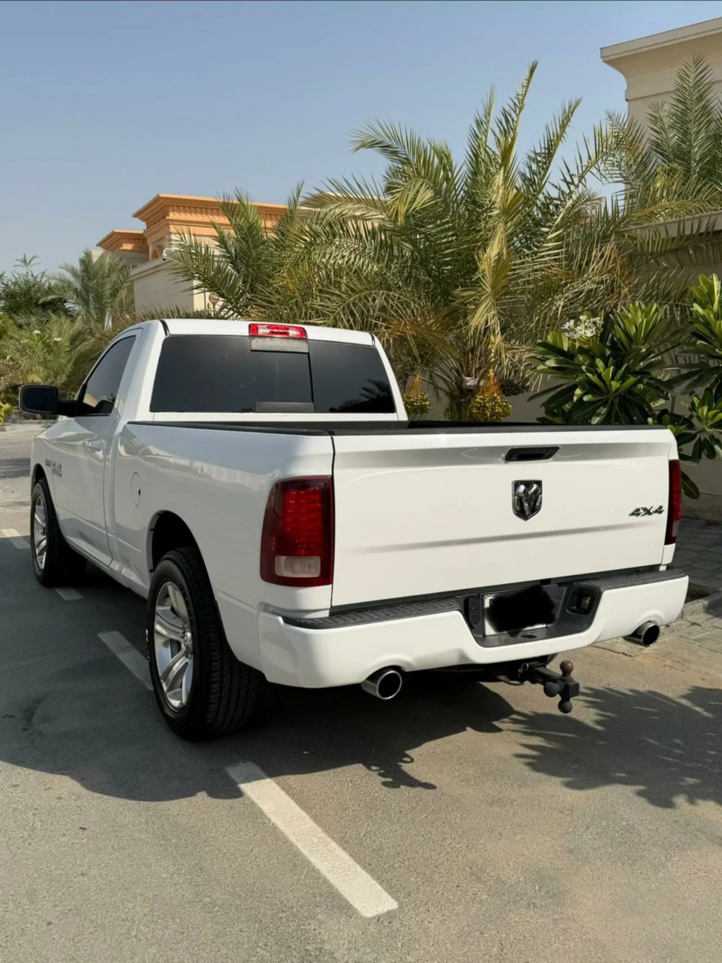 White pickup truck parked in a parking lot. The truck is a Dodge Ram, with a black bed liner and a red tailgate. It has a