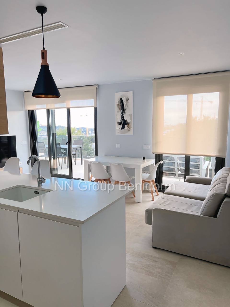 Modern and spacious living room and kitchen area. The room has a large window with white blinds, allowing natural light to