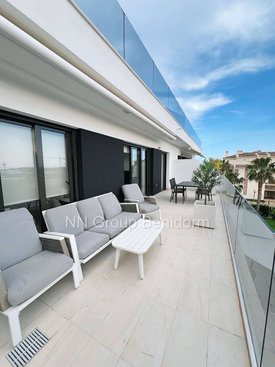 Modern balcony with a white tiled floor and a glass railing. The balcony has a gray sofa set with two armchairs and a small