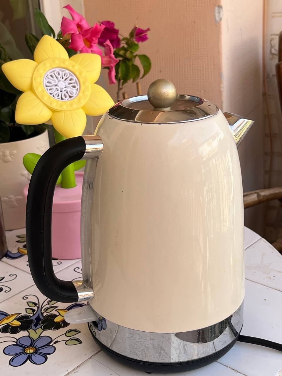 White electric kettle with a black handle and a silver lid. The kettle is placed on a white table with a floral patterned