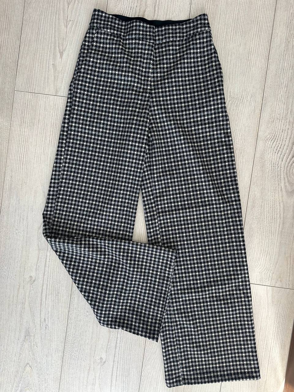 Pair of black and white houndstooth trousers lying on a wooden floor. The trousers have a high waist and wide legs, with a