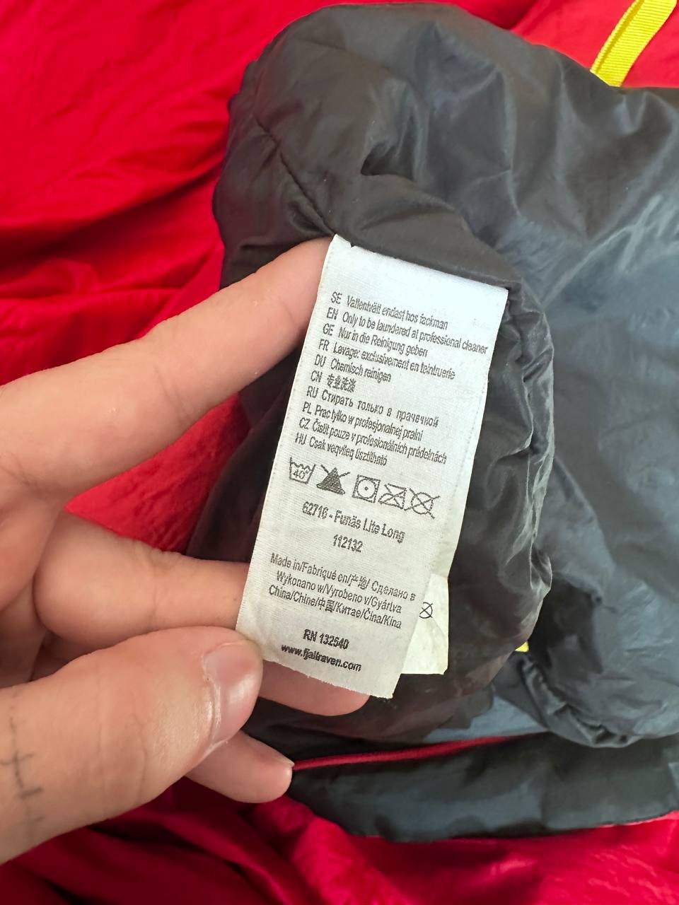 Hand holding a black sleeping bag with a white label attached to it. The label has text written on it in black ink and