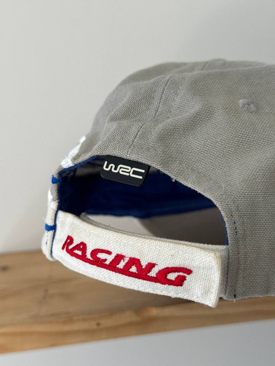 Close-up of a baseball cap with the word "RACING" written in red and white on the front. The cap is made of a light grey