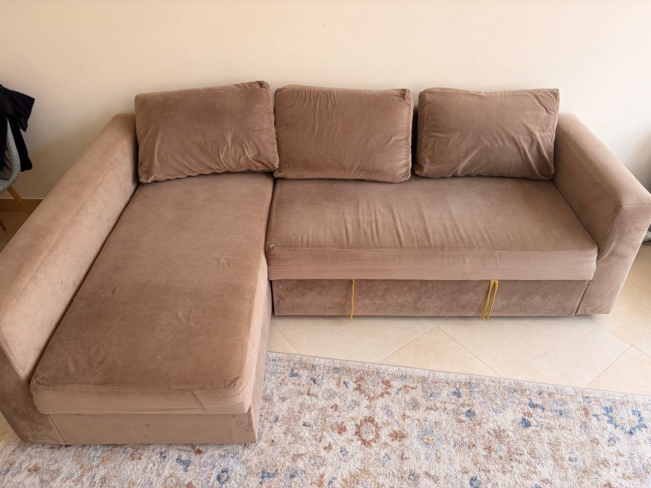 Beige-colored sofa in a living room. The sofa has a chaise lounger on the left side and a small ottoman on the right side.