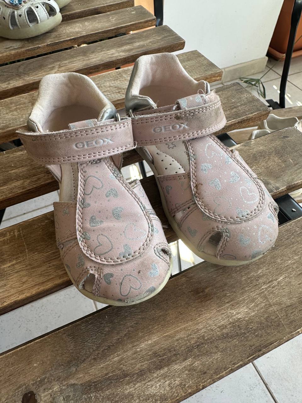 Pair of beige-colored baby shoes on a wooden bench. The shoes have a velcro strap across the top of the foot and a small