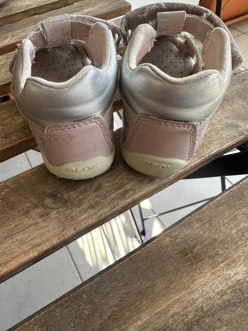 Pair of baby shoes on a wooden bench. The shoes are light pink in color and have a velcro strap across the top of the foot.