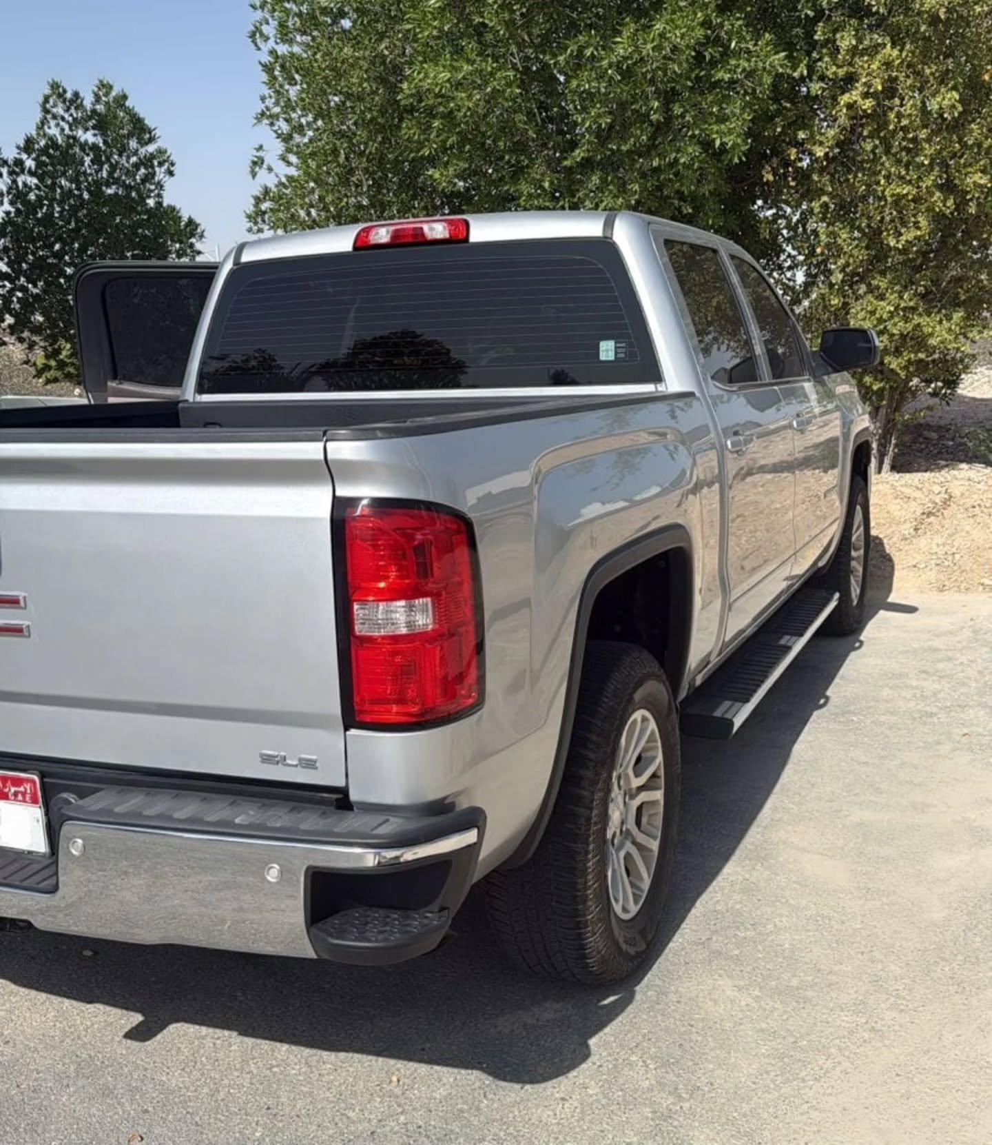 Silver pickup truck parked on a concrete driveway. The truck has a black bed liner and a red tail light. The bed liner is