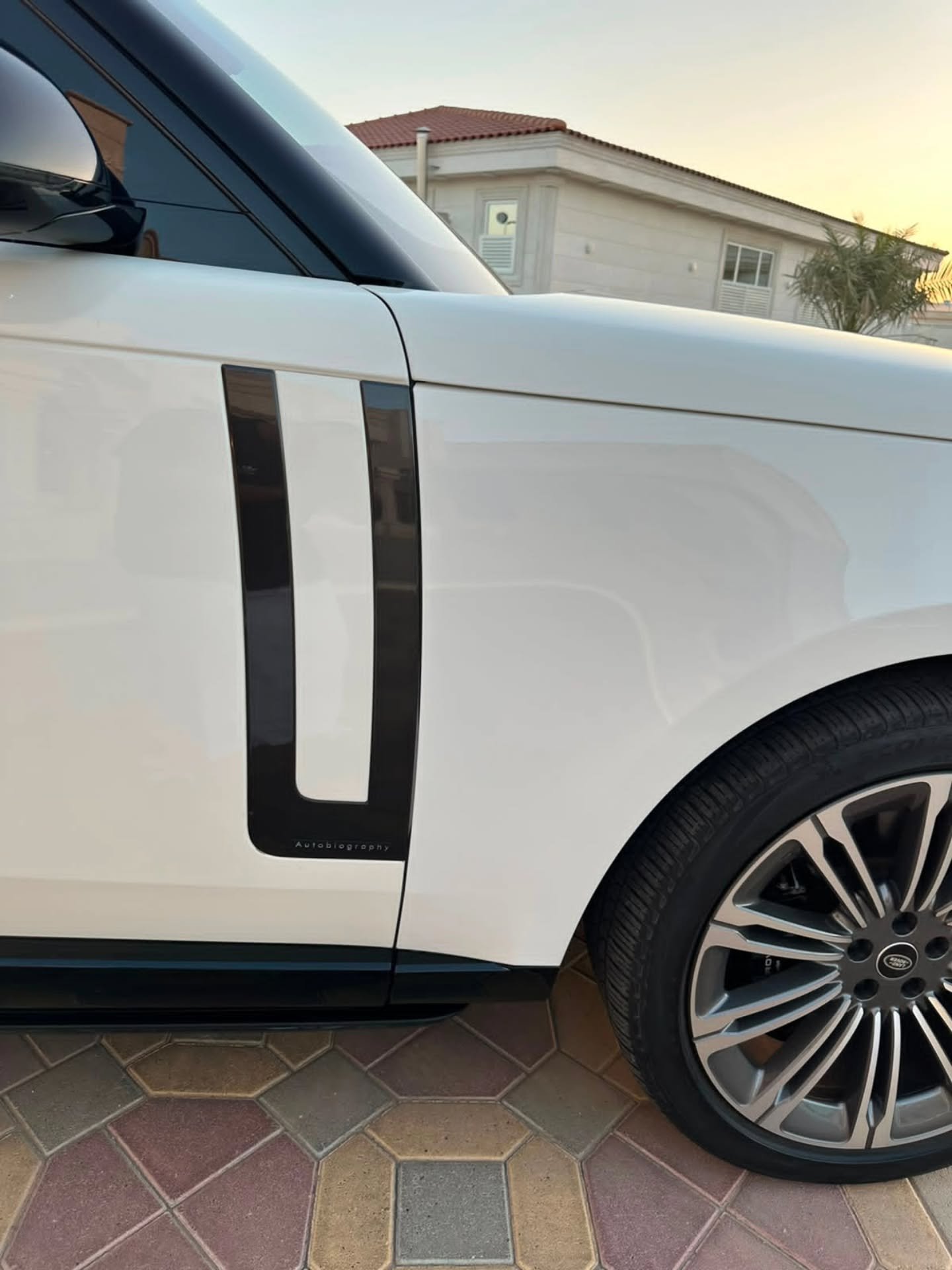 White car parked on a tiled driveway. The car has a black side stripe on the side and a silver alloy wheel. The background