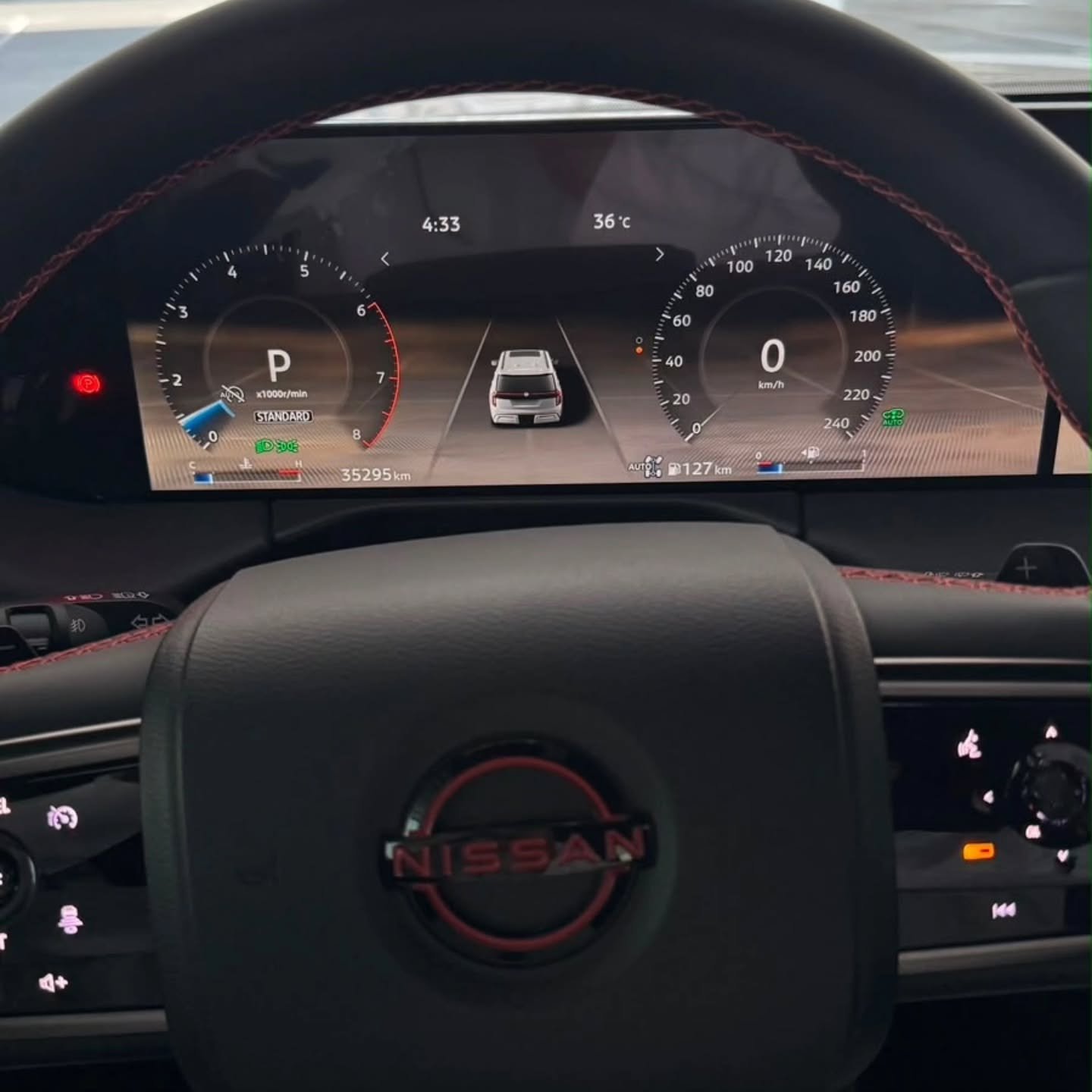 Dashboard of a car, specifically the steering wheel and the Nissan logo. The steering wheel is black with red stitching and