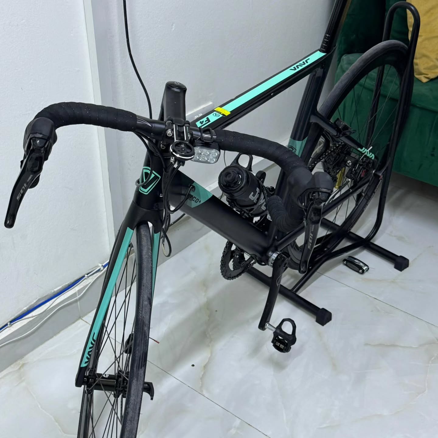 Black and green bicycle with a black frame and handlebars. The bike is parked on a white tiled floor in a room with a green