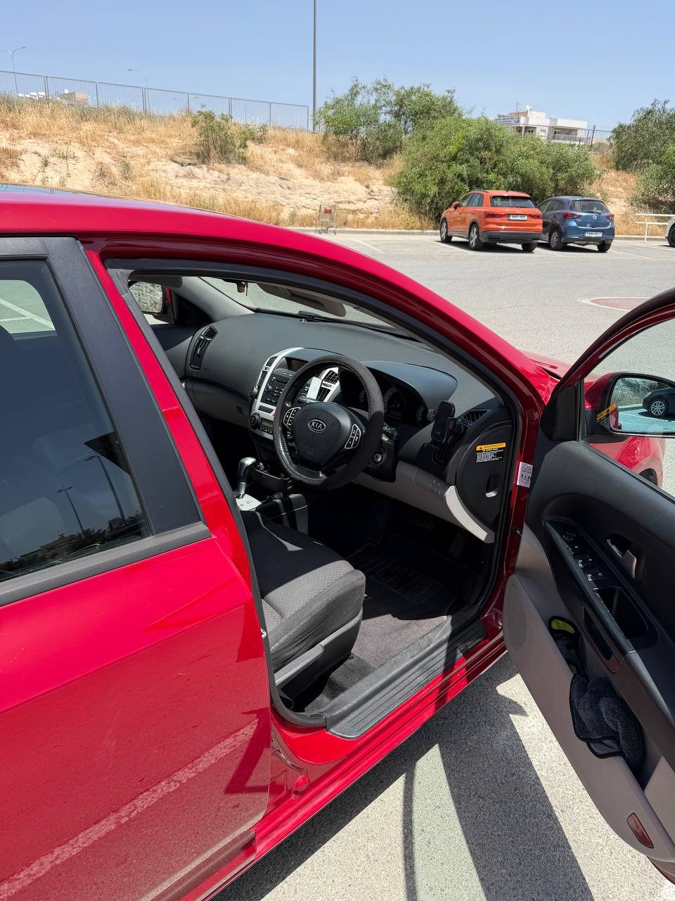 Interior of a red car with the door open. The car is parked in a parking lot with other cars visible in the background. The