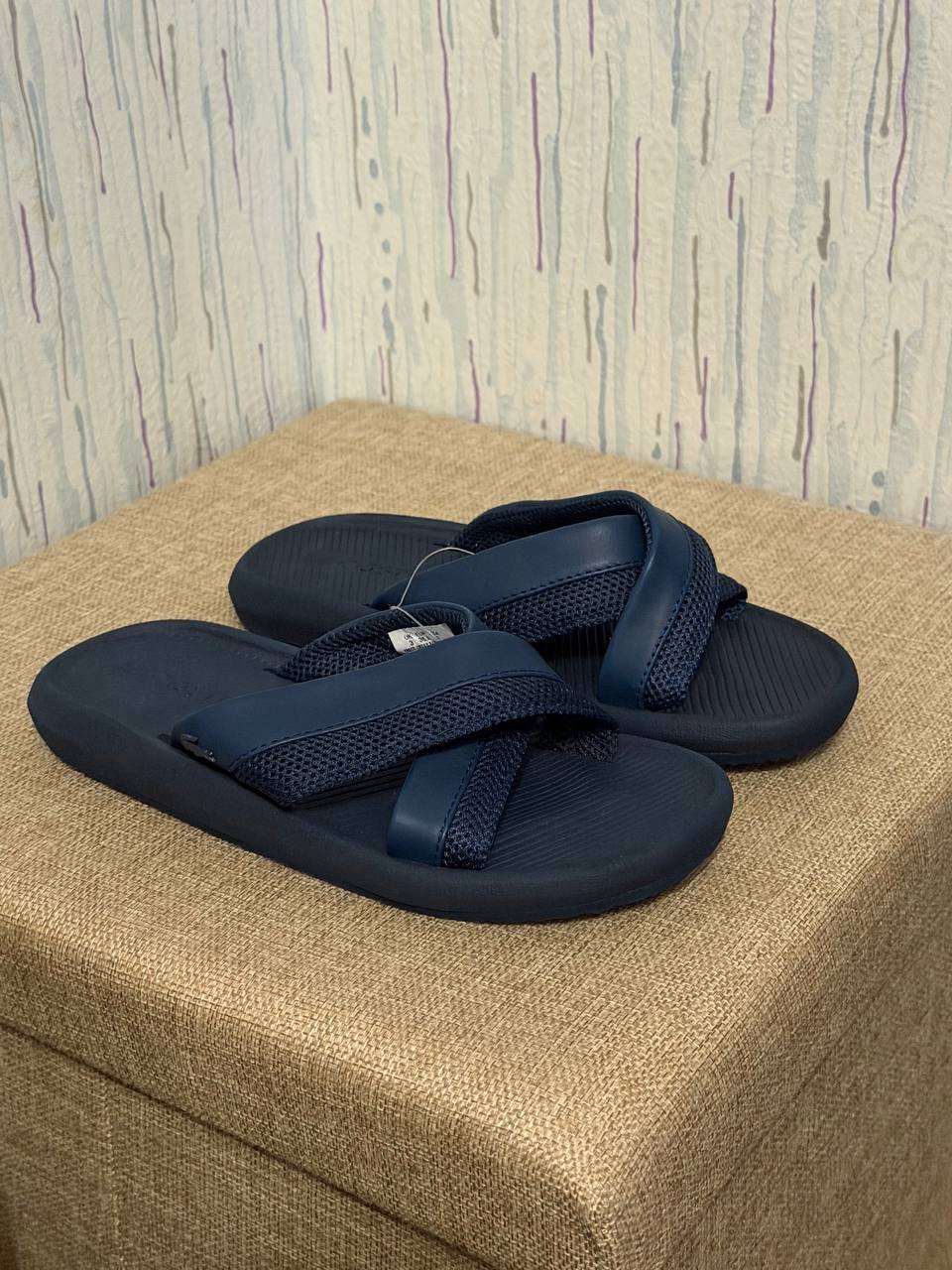 Pair of navy blue flip-flops resting on a beige-colored ottoman. The slippers have a strap across the top of the foot with a