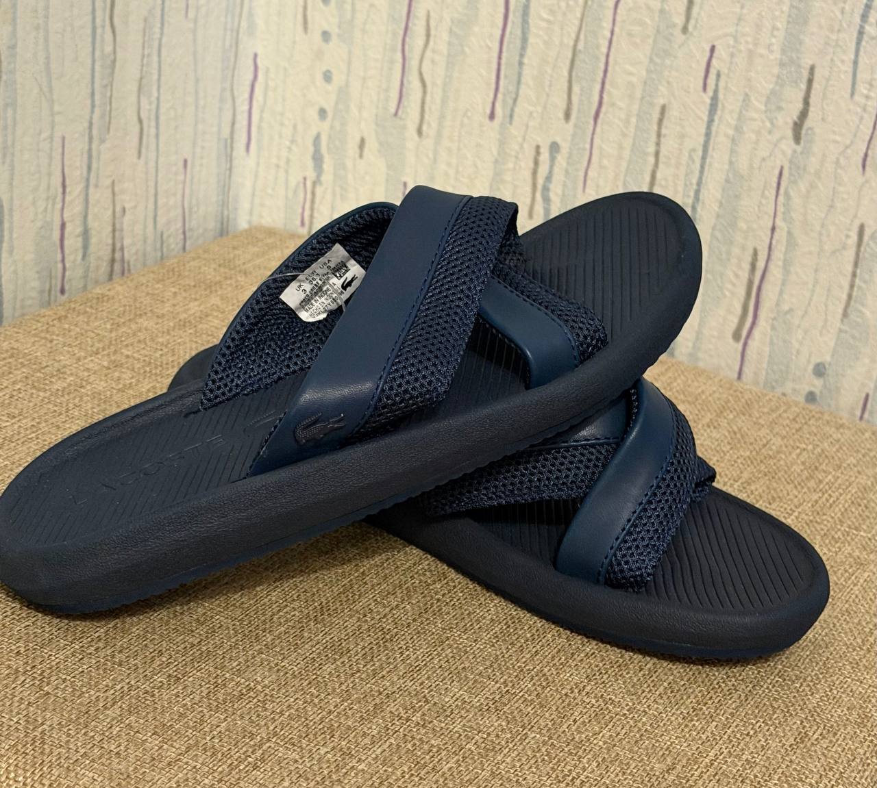 Pair of navy blue flip-flops on a beige surface. The sandals have a strap across the top of the foot with a small white tag