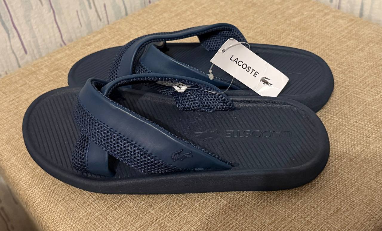 Pair of navy blue Lacoste flip-flops on a beige couch. The sandals have a strap across the top of the foot with the brand's