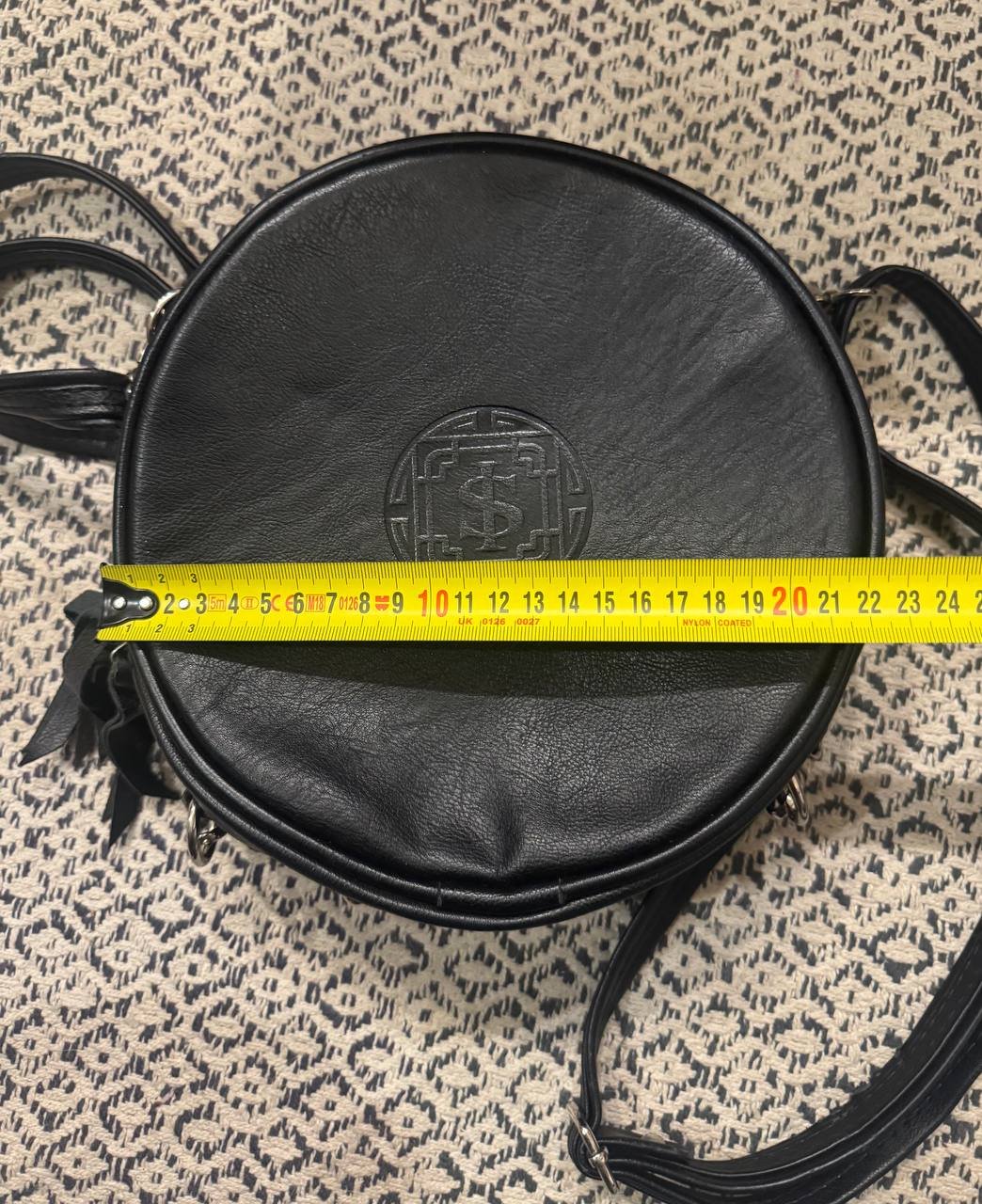 Black round leather bag with a gold logo on the front. The bag has two black straps on either side and a yellow measuring