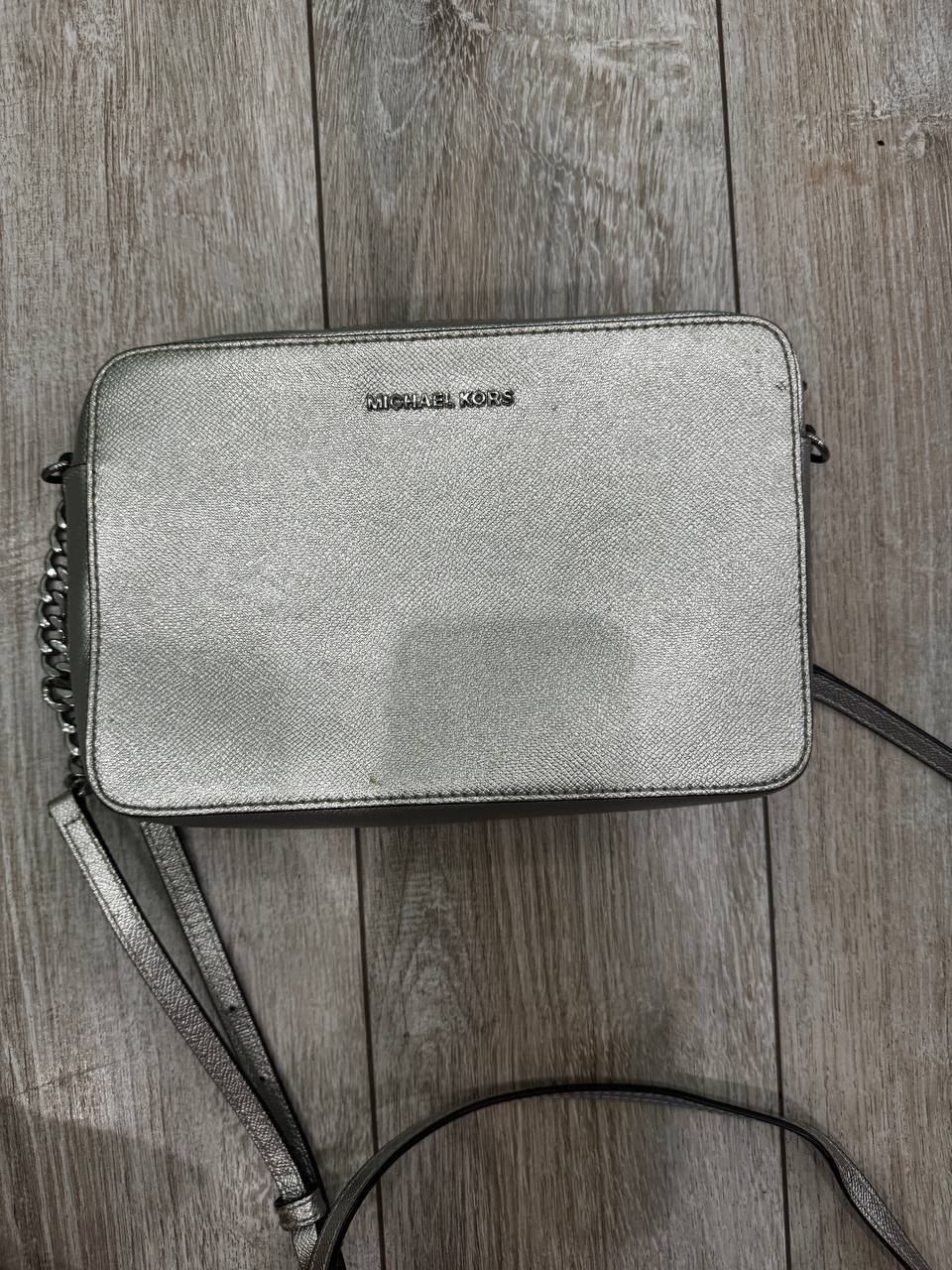 Silver crossbody bag with a chain strap. The bag is made of a textured, metallic material and has the brand name "Michael