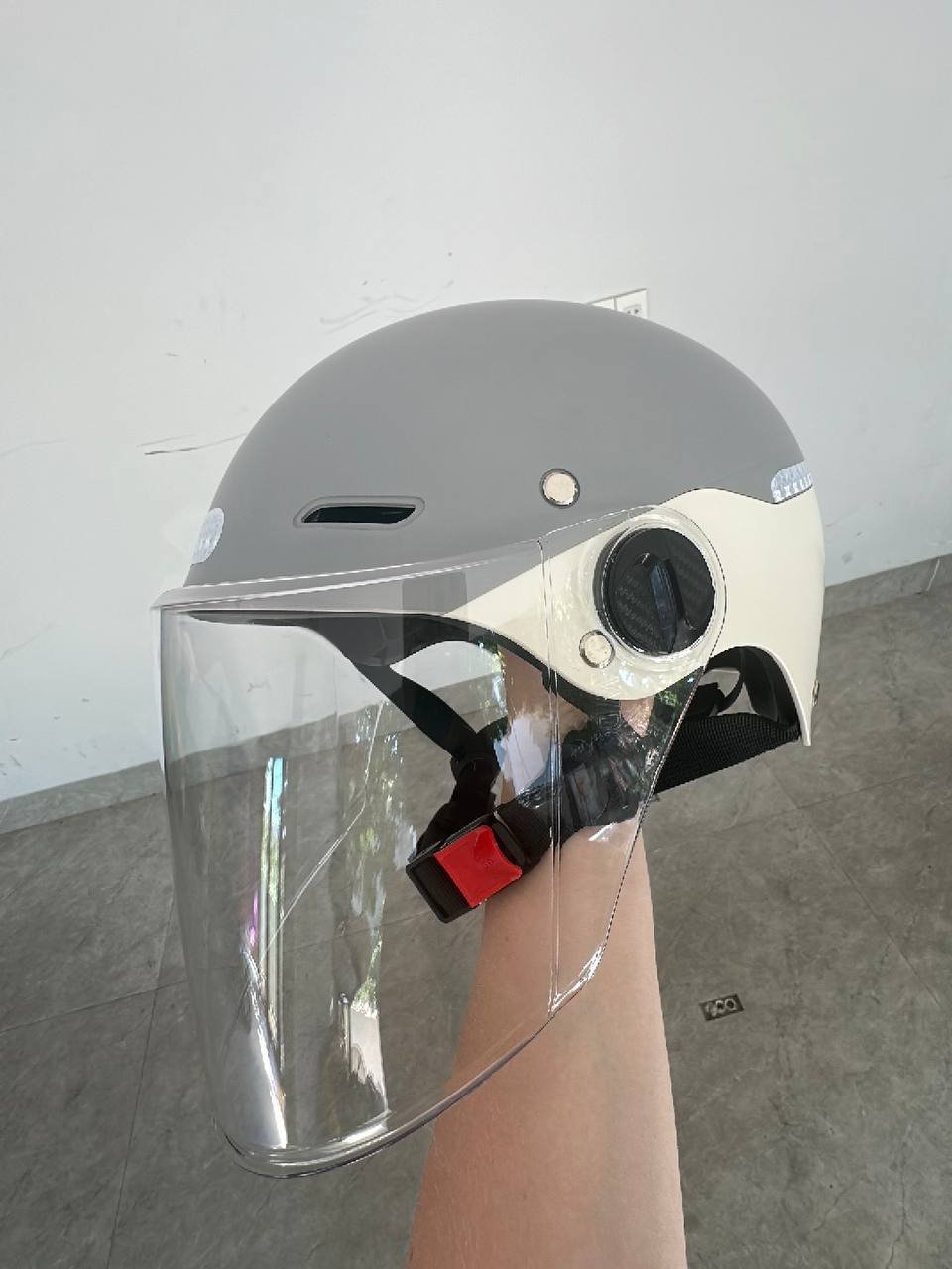 Person's hand holding a helmet with a clear visor. The helmet is grey in color and has a white visor on the front. The visor