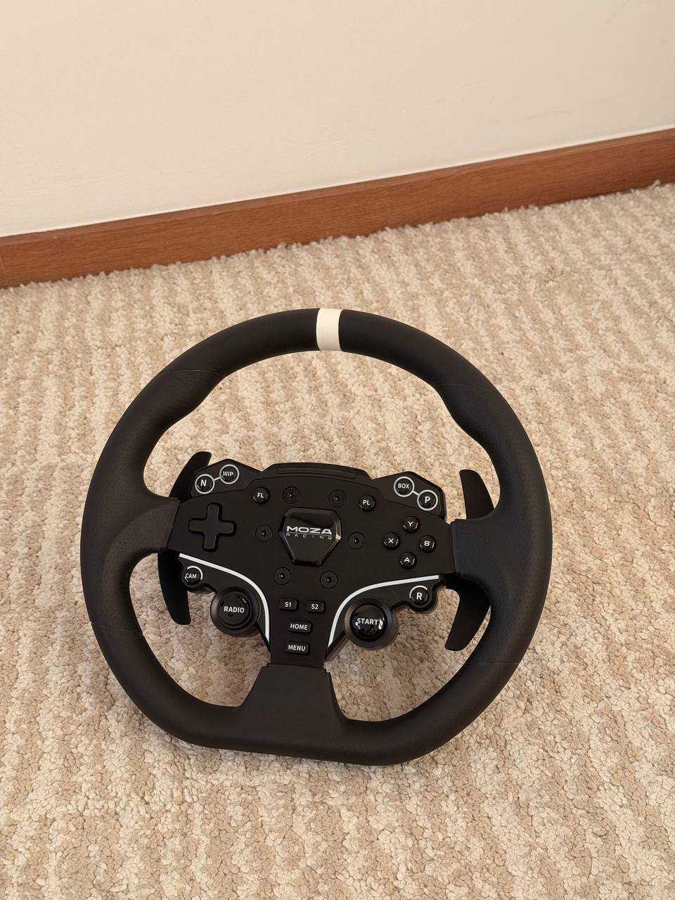 Black steering wheel with a white stripe on the top. The steering wheel is resting on a beige carpeted floor. It has a black