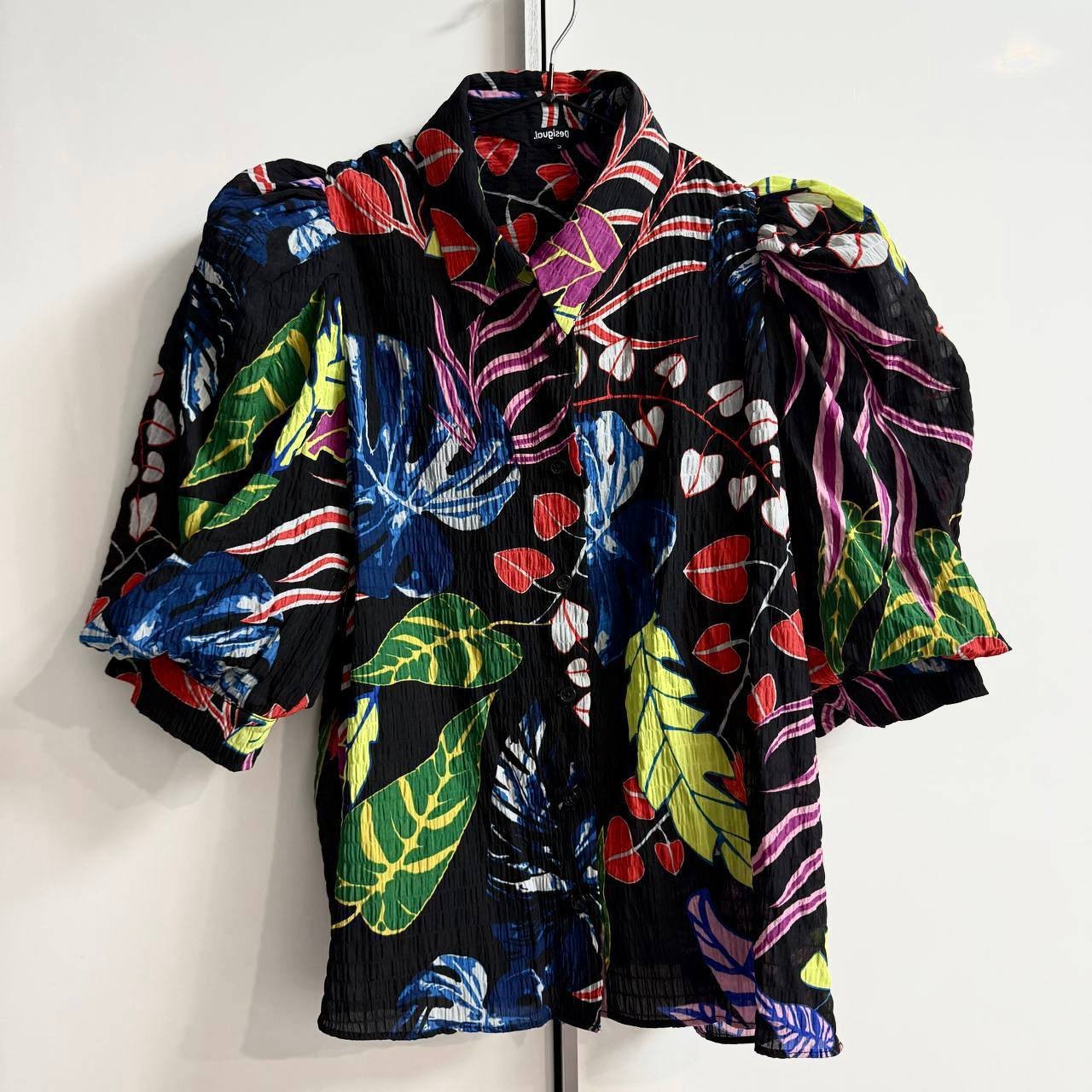 Black blouse hanging on a hanger against a white wall. The blouse has a collared neckline and short sleeves. It is made of a