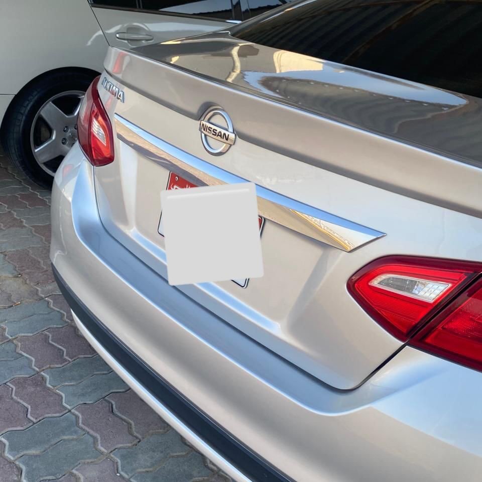 Rear end of a silver Nissan car. The car is parked on a cobblestone street with other cars visible in the background. The