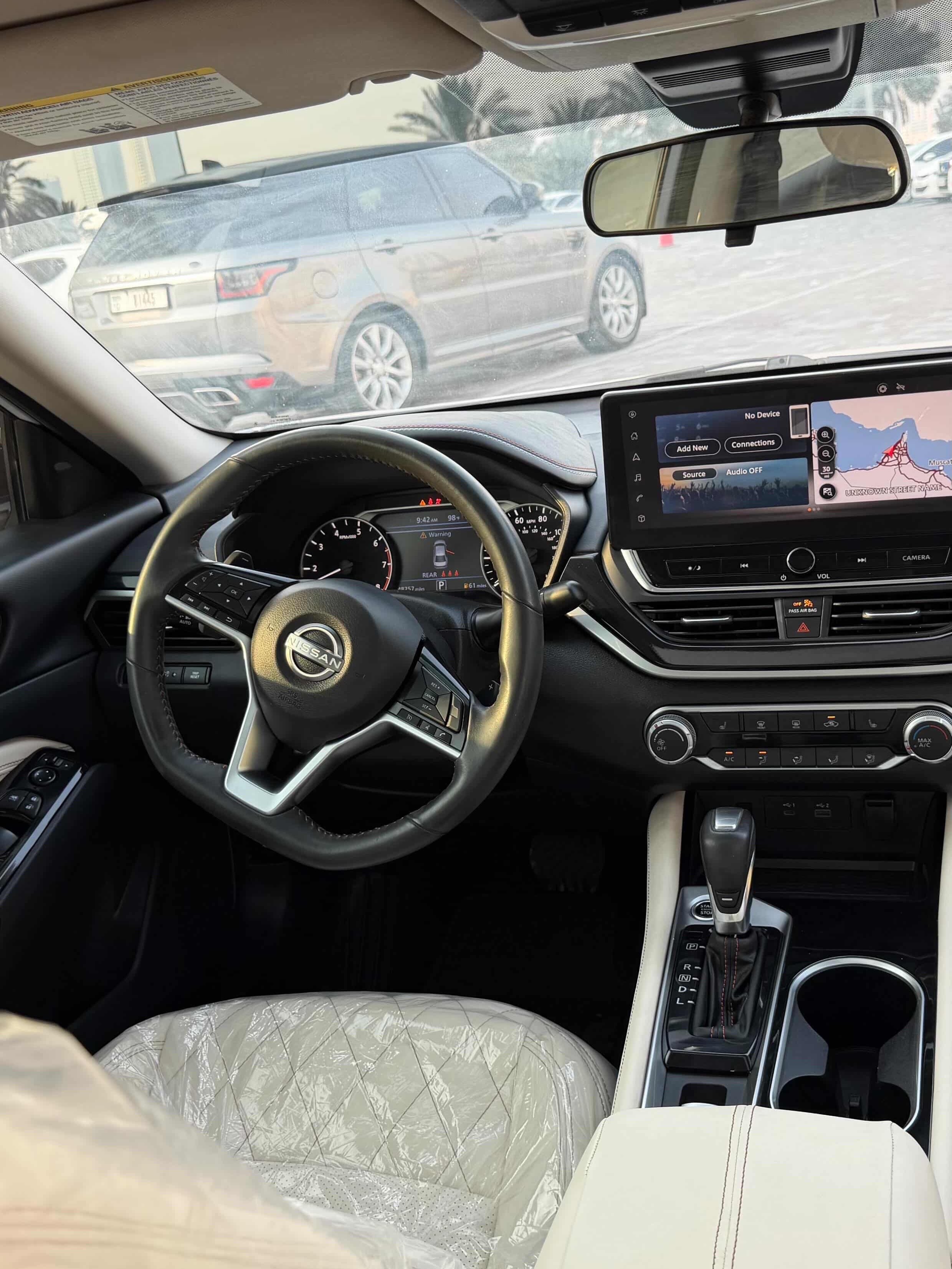 Interior of a car, specifically the driver's seat and dashboard. The car appears to be a Nissan, as the logo of the car is