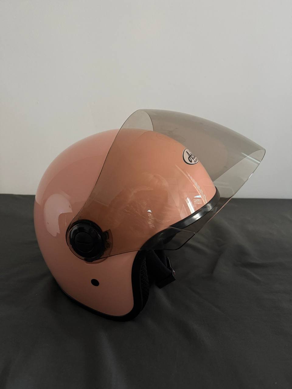 Pink helmet with a clear visor. The helmet is resting on a black surface with a white wall in the background. The visor is