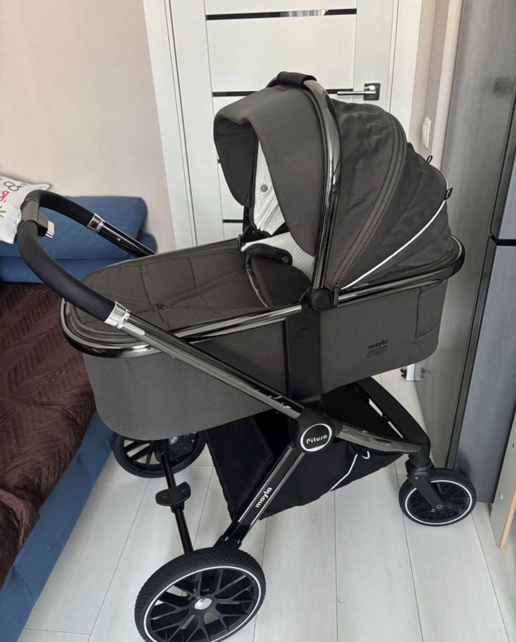 Baby stroller in a room with a white door and a blue couch in the background. The stroller is black and grey in color and