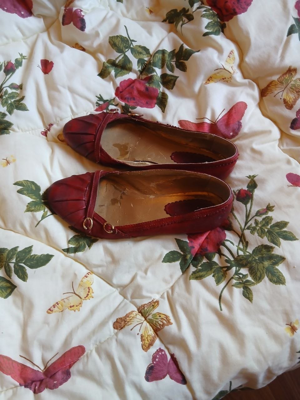 Pair of red flats lying on a white bedsheet with a floral pattern. The flats have a pointed toe and a low heel. The fabric