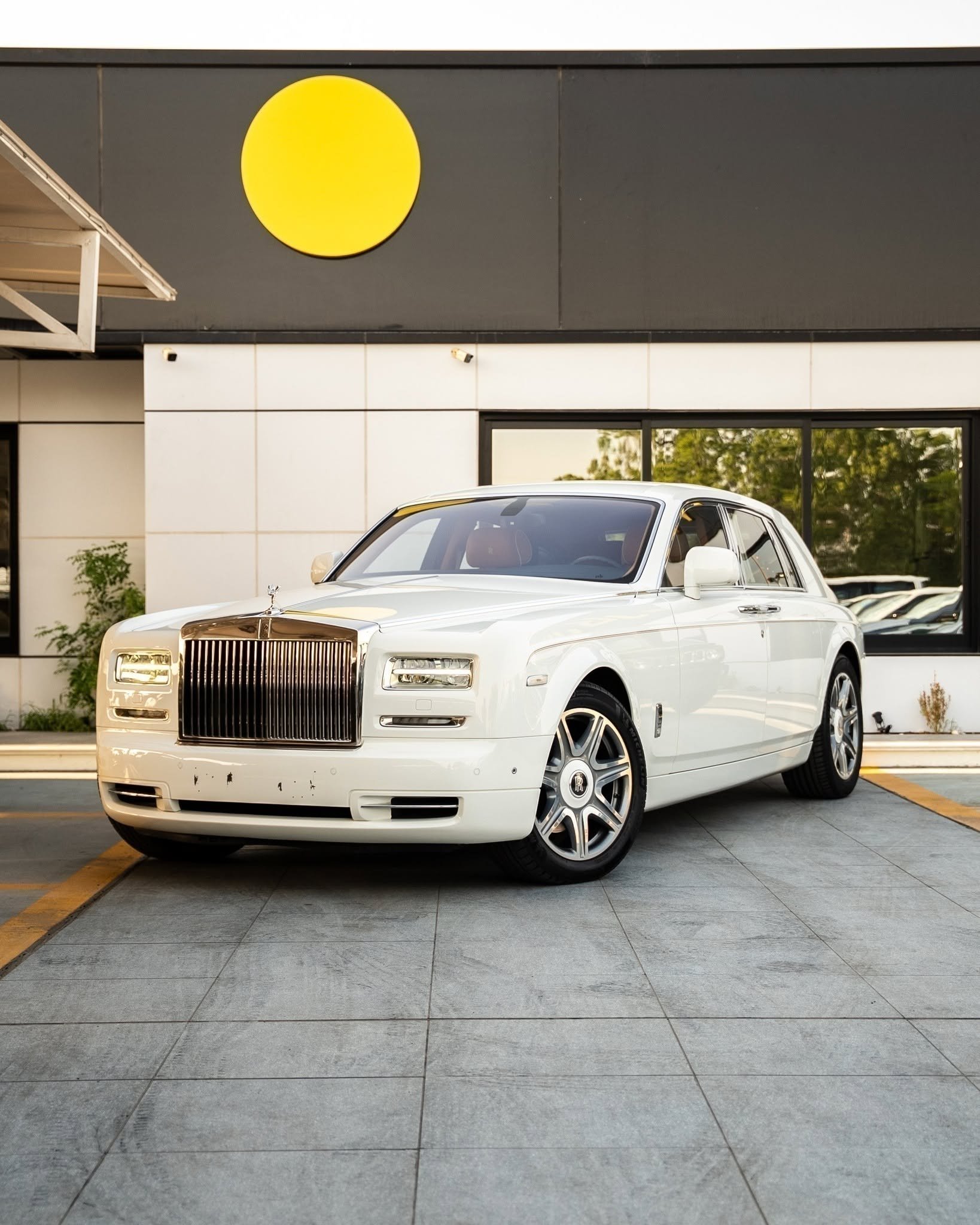 White Rolls Royce car parked in front of a modern building with a large yellow circle on the wall. The car is a luxury model