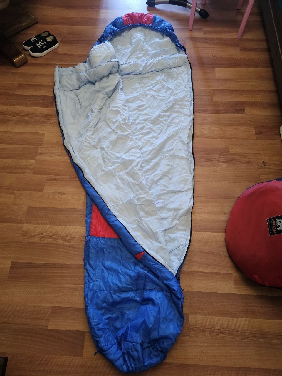 Blue and white sleeping bag lying on a wooden floor. The sleeping bag is rectangular in shape and appears to be made of a