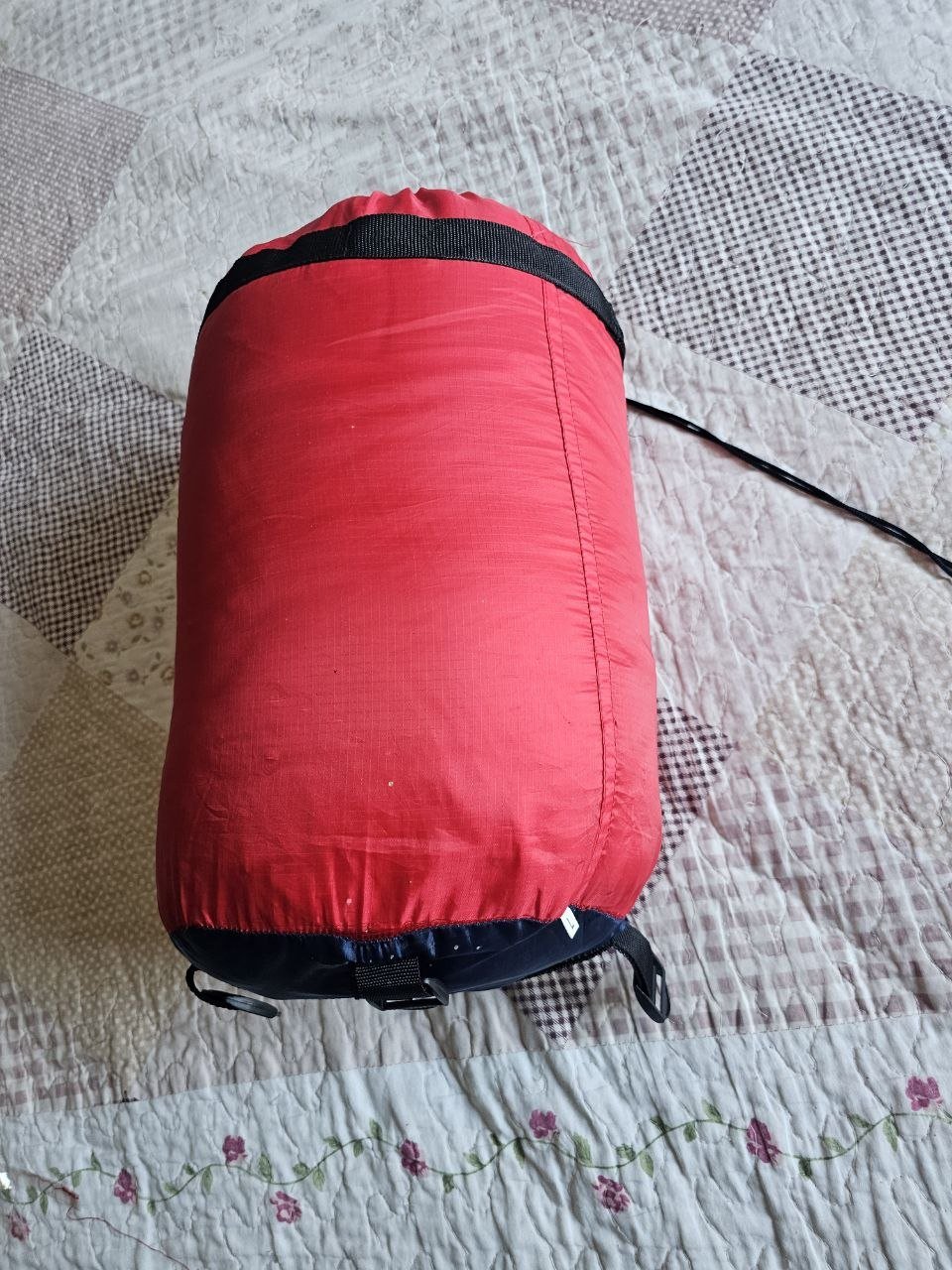 Red sleeping bag lying on a white quilt with a floral pattern. The sleeping bag is cylindrical in shape and appears to be