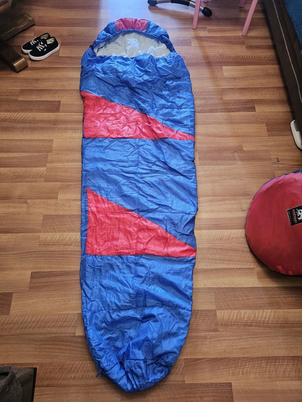 Blue sleeping bag lying on a wooden floor. The sleeping bag is rectangular in shape and has a red and blue striped pattern