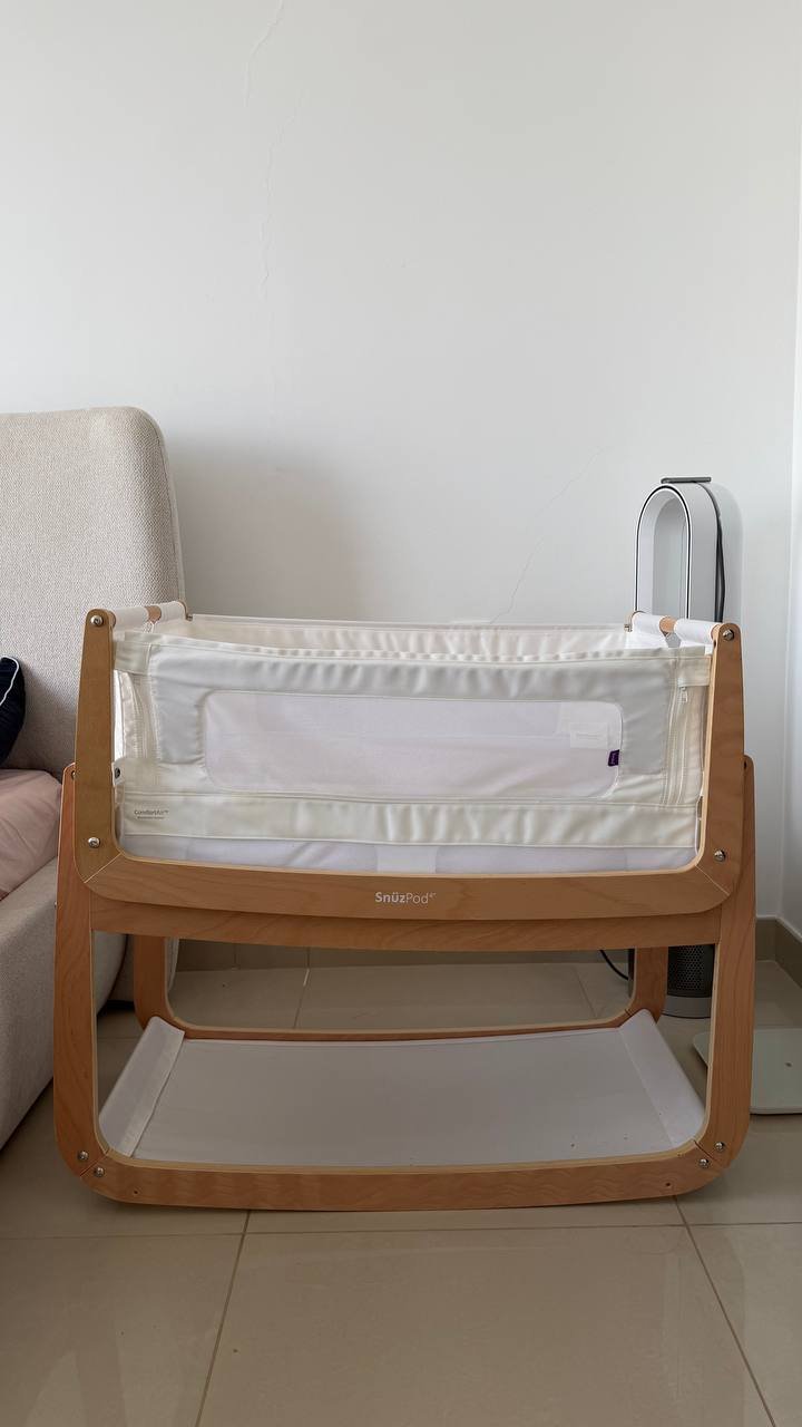 Wooden baby bassinet in a corner of a room. The bassinet is made of light-colored wood and has a white mattress on top. The