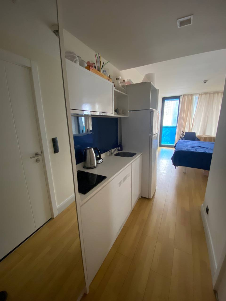Small kitchen and bedroom in a small apartment. The kitchen has white cabinets and a white refrigerator and sink. There is a