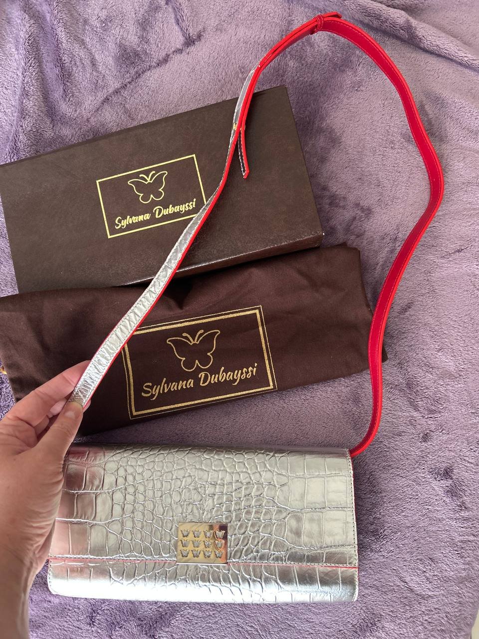 Hand holding a silver clutch bag with a red strap. The bag is made of a crocodile-embossed leather material and has a